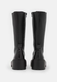 Botas negras hasta la rodilla, hechas de cuero liso, con suela gruesa, costuras verticales, cierre con cremallera y una etiqueta con el logo en la parte posterior.