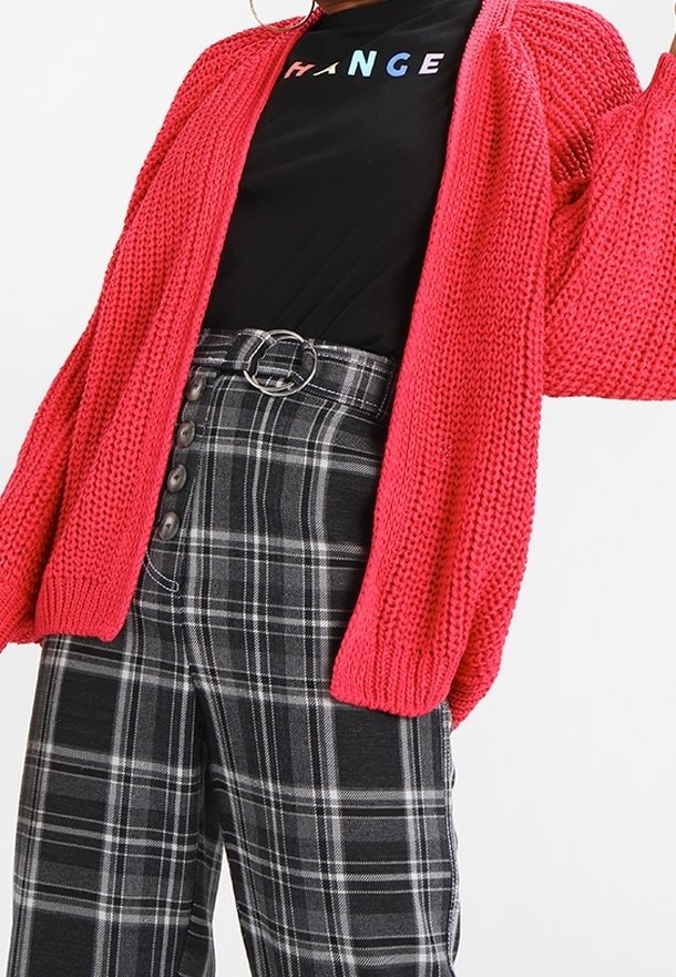 Chemise noire avec texte multicolore « CHANGE », cardigan rouge en grosse maille, et pantalon taille haute à carreaux noirs et gris avec détails de boutons et ceinture à anneau.