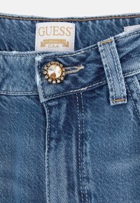 Dżinsy w jasnoniebieskim kolorze, z błyszczącym, ozdobnym guzikiem i złotą metką. Widoczne szycie i gładka tekstura w całej powierzchni.