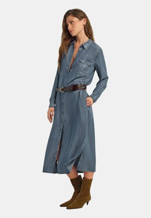 Donna che indossa un vestito camicia di denim blu a maniche lunghe con una cintura marrone e stivali marroni in camoscio con tacco a metà polpaccio, in piedi contro uno sfondo semplice.