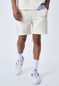 Pantalones cortos de color crema con un ajuste relajado, que presentan bolsillos laterales, un acento en lila y detalles bordados en la parte delantera. Acabados con una textura suave.