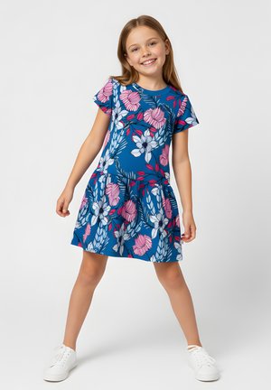 Uśmiechnięta dziewczyna stojąca z szeroko rozstawionymi nogami, ubrana w niebieską sukienkę z dużymi różowymi i białymi kwiatowymi wzorami oraz białe trampki.