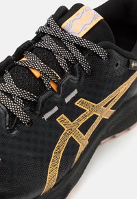 Črne atletske čevlje z mrežasto zgornjim delom z teksturo, z zlatim logotipom ASICS, črno-bež potiskanimi vezalkami in oranžnimi poudarki na ježku.