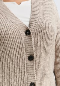 Cardigan en tricot beige avec un décolleté en V, texture côtelée et cinq boutons noirs sur le devant. Le tissu semble doux et chaud.