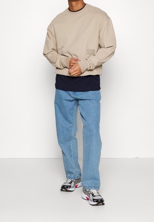 Beige sweatshirt with a crew neck, front pocket, paired with a navy shirt, light blue jeans, and black/red athletic sneakers.