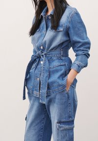 Chaqueta de mezclilla con cinturón en la cintura, dos bolsillos en el pecho y cierre de botones. La tela es de un lavado azul intermedio, con una textura ligeramente usada.