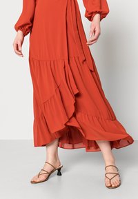 Czerwona warstwowa sukienka z falbanami, długimi rękawami i przewiązaniem z boku. W zestawie z brązowymi sandałami na obcasie z paskami. Gładka tekstura tkaniny.