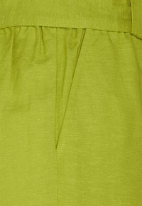 Zelená látka s hladkým texturem, s nařasenou částí nad boční kapsou. Design zdůrazňuje jednoduchost a funkčnost.