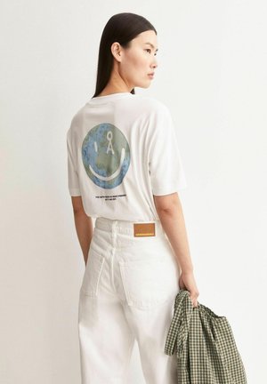 Woman wearing white pants and a white T-shirt with a blue and green earth smiley graphic and text on the back, holding a green plaid shirt.