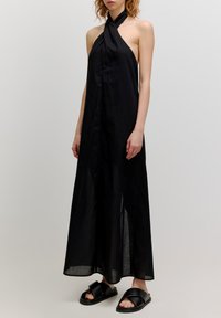 Zwarte halter maxi-jurk van lichte stof met een gedraaide halslijn en een losvallende, vloeiende silhouet. Gedragen met zwarte platte schoenen.