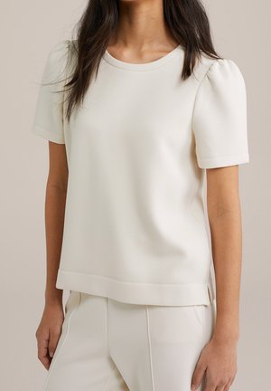 Vrouw die een crèmekleurige top met korte mouwen en opgeblazen schouders draagt, gecombineerd met crèmekleurige broek, staand tegen een neutrale achtergrond.