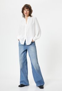 Hvid langærmet bluse med knapfront og en let glans, parret med brede, falmede blå denimjeans og sorte firkantede sko.