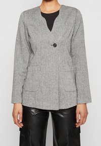 Blazer texturé gris avec un col en V, une fermeture à un bouton noir unique et deux poches avant. Associé à un pantalon noir en simili cuir.