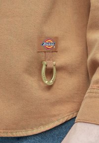 Καφέ ύφασμα με υφή, που διαθέτει μεγάλο χρυσό μεταλλικό δαχτυλίδι σε σχήμα πεταλούδας, προσαρτημένο σε καφέ ταμπέλα με το λογότυπο Dickies.