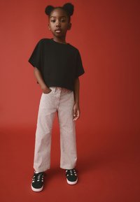 Schwarzes kurzärmeliges Oberteil, rot-weiß gestreifte Hosen mit weitem Bein und schwarze Sneakers mit weißen Akzenten vor rotem Hintergrund.