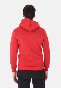 Uomo in piedi di spalle, che indossa un hoodie rosso con il cappuccio abbassato e pantaloni neri, davanti a uno sfondo chiaro e semplice.