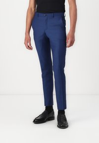 Pantalon bleu sur mesure en tissu lisse, avec une coupe ajustée, un devant plat et des poches latérales. Associé à des mocassins noirs et des chaussettes.
