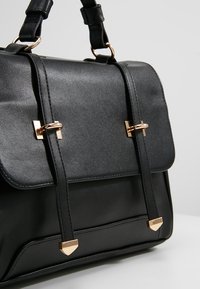 Sac à main en cuir noir avec deux sangles avant sécurisées par des fermoirs métalliques dorés et une quincaillerie assortie dorée sur les poignées.