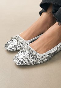 Ezüst flitterekkel borított balettcipő fehér szegéllyel és díszes masnival az elején. Lapos talp és lekerekített orr kialakítás.