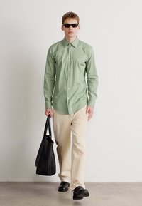 Homem com camisa verde de botões, calças bege, sapatos pretos, óculos de sol, carregando uma grande bolsa preta, caminhando no interior contra uma parede branca.