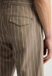Gestreifte Hosen in Olivgrün mit dünnen roten Streifen, strukturiertem Stoff und einer einzelnen Gesäßtasche mit metallischem Druckknopfverschluss.