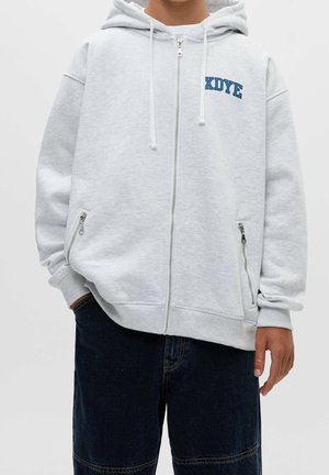 Persona que lleva una sudadera con capucha y cremallera en gris claro con el texto azul "XDYR" en el pecho, manos en los bolsillos, combinada con pantalones cortos de mezclilla azul oscuro.
