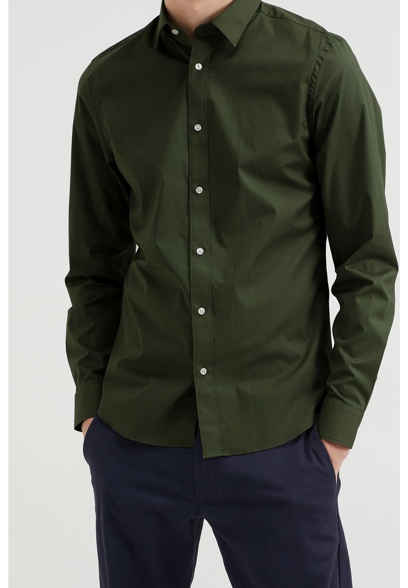 Mörkgrön skjorta med knappar, långa ärmar, lätt textur, med silverknappar och klassisk krage, bärs med marinblå byxor.