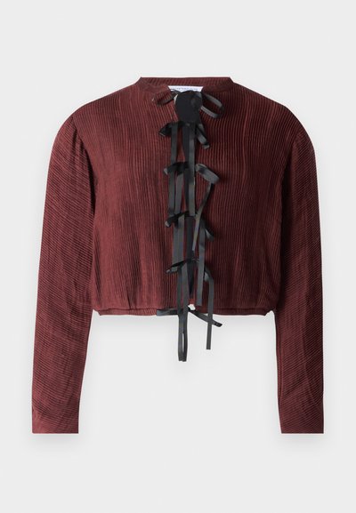 Kastanjas sarkans īsais tops, kas izgatavots no teksturēta auduma. Tam ir garas piedurknes un priekšējais aizdare ar melniem lentes aizdariem, kas sakārtoti vertikāli.