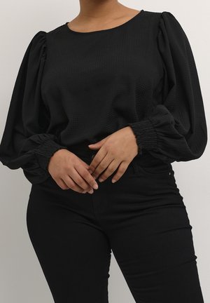 Vrouw draagt een zwarte blouse met structuur, pofmouwen en geplooide manchetten, gecombineerd met een hoge zwarte broek, handen voor zich in elkaar gevouwen.