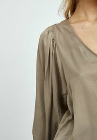 Beige Bluse mit tiefem V-Ausschnitt, langen gerafften Ärmeln und glatter Satinoberfläche. Der Stoff liegt weich am Körper an.
