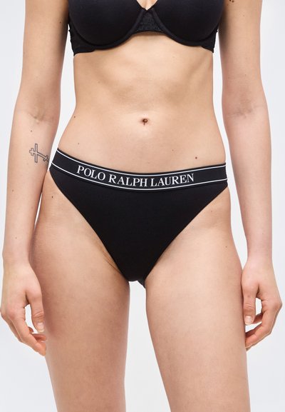Γυναίκα που φοράει μαύρο ψηλοκάβαλο εσώρουχο με ζώνη μέσης με την επωνυμία "Polo Ralph Lauren" και ένα αντίστοιχο μαύρο σουτιέν, στέκεται μπροστά σε λευκό φόντο.