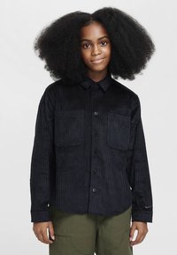 Czarna koszula sztruksowa z długimi rękawami, zapinana na guziki z przodu, z dwoma kieszeniami na piersi i miękką fakturą. Na rękawie znajduje się subtelne logo.