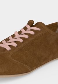 Καφέ σουέτ αθλητικό παπούτσι με ανοιχτό ροζ κορδόνια και μπεζ σόλα, απεικονίζεται σε κοντινή γωνιακή θέα.