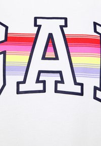 T-shirt bianco in cotone con grandi lettere in grassetto "GA" contornate di blu navy, accentuate da strisce orizzontali in rosso, rosa, giallo e viola.