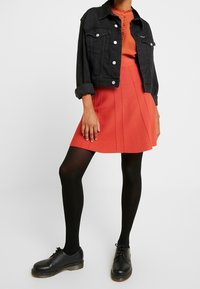 Schwarze Denim-Cropped-Jacke, orangefarbener Rüschentop, roter A-Linien-Rock, schwarze Strumpfhosen und schwarze Schuhe mit beiger Sohle. Strukturierter Schnitt und Passform.