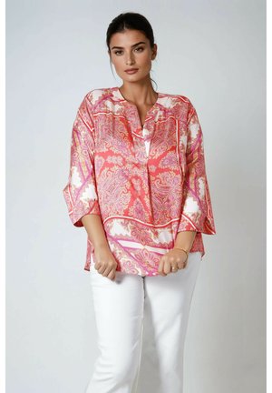 Femme portant un chemisier ample à manches longues rose et blanc à motif cachemire, avec un pantalon blanc, debout devant un fond gris clair uni.
