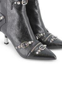 Botas de tobillo de imitación de cuero negras con punteras puntiagudas, remaches plateados, cremalleras y detalles de hebilla. Presentan un diseño de tacón metálico bajo.