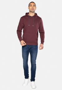 Bordeaux hoodie met trekkoorden, kangoeroezak, gedragen met blauwe jeans en witte sneakers. Zachte stof, geribde manchetten en zoom.
