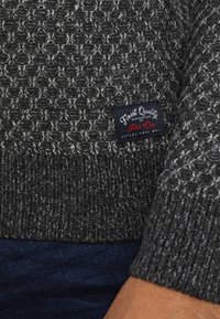 Koolgrijs gebreid vest met een textuurpatroon en geribbelde zoom. Voorzien van een zwart etiket met witte en rode tekst, waarop staat "Finest Quality."
