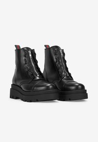 Botas de cuero negras hasta el tobillo con una suela gruesa, cremallera frontal y detalles de cordones. Presentan una lengüeta de tirón con rayas verde y roja en el tobillo.