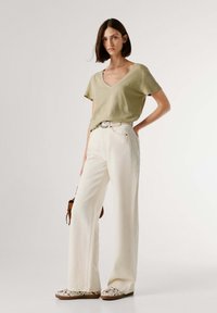 Γυναίκα που στέκεται φορώντας ανοιχτό πράσινο T-shirt με V λαιμόκοψη, παντελόνι με φαρδιά πόδια και ψηλή μέση σε εκρού χρώμα, μοκασίνια με σχέδια και κρατώντας μια μικρή καφέ τσάντα.