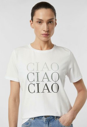 Kobieta o ciemnych włosach w białej koszulce z napisem "CIAO" powtarzającym się trzykrotnie w literach z gradientowych kryształków, w połączeniu z niebieskimi dżinsami.