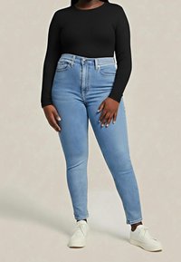 Femme portant un jean skinny taille haute bleu clair, un haut noir à manches longues et des baskets blanches, debout sur un fond uni.