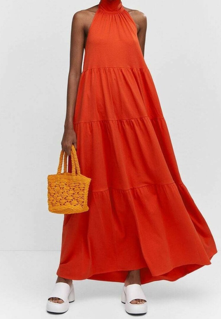 Femme portant une robe longue à volants orange vif sans manches, tenant un petit sac à main en tissage orange et des sandales compensées blanches.