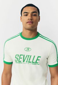Bílý tričko s zelenými akcenty, na kterém je vytištěno „SEVILLE“ a „ESPAÑA“ tučným, opotřebovaným písmem. Kulatý výstřih a krátké rukávy.