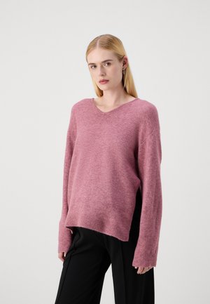 Pull en tricot rose avec un col en V et des manches longues légèrement oversize. Texture douce et ourlet côtelé. Associé à un pantalon noir.