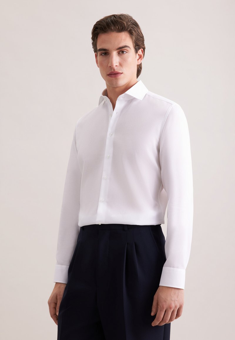 Chemise blanche boutonnée avec col, manches longues et texture lisse, associée à un pantalon bleu marine foncé.