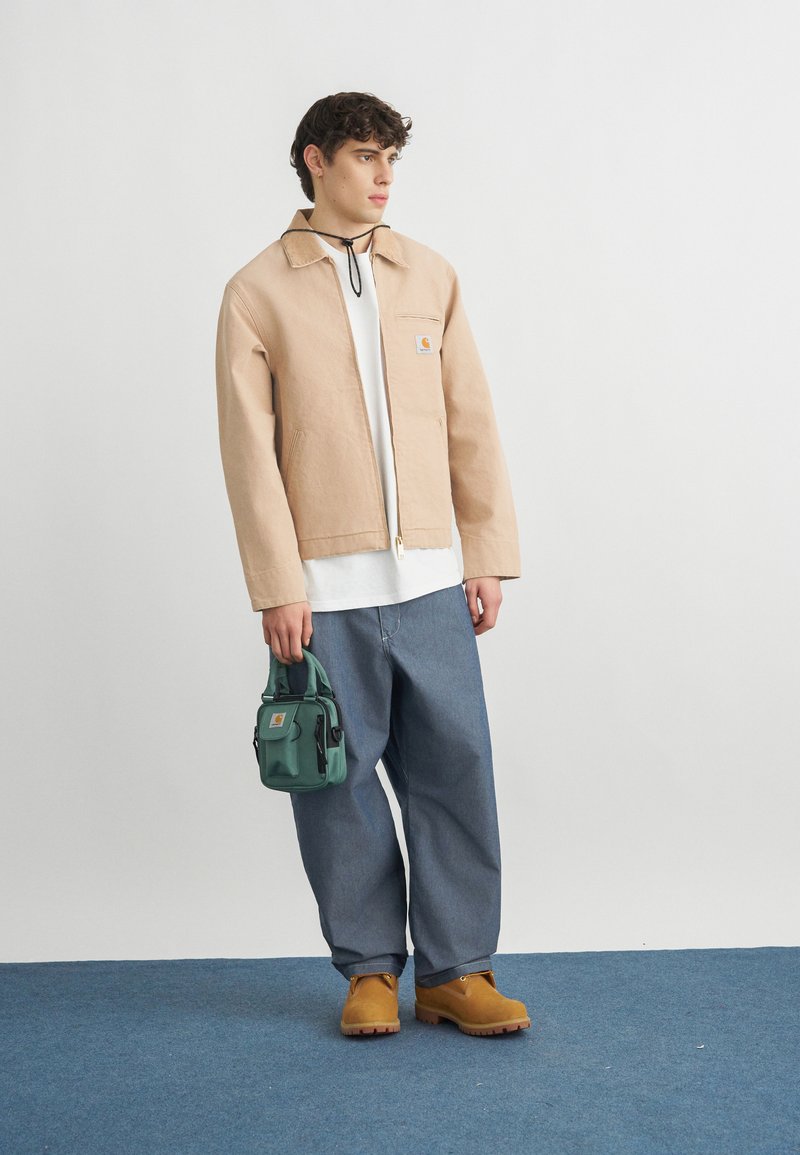 Giovane uomo con giacca beige, camicia bianca, jeans blu larghi e stivali marroni, che tiene una piccola borsa verde, in piedi su un tappeto blu.