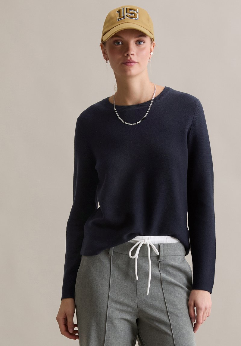 Marineblauer Pullover mit langen Ärmeln, weiche Textur. Kombiniert mit hellgrauer Hose und weißem Kordelzug. Trägt eine beige Mütze mit der Nummer 15.