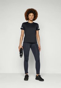 Camiseta atlética preta de mangas curtas com logo na manga, combinada com leggings escuras e ténis pretos. Garrafa de água segurada na mão.